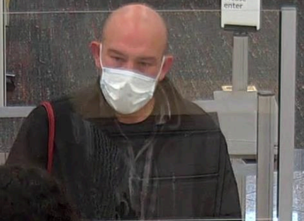 White, bald male who is 40-50 years old. He is wearing a surgical mask, an over-sized dark coat, and a striped shirt over a dark t-shirt.